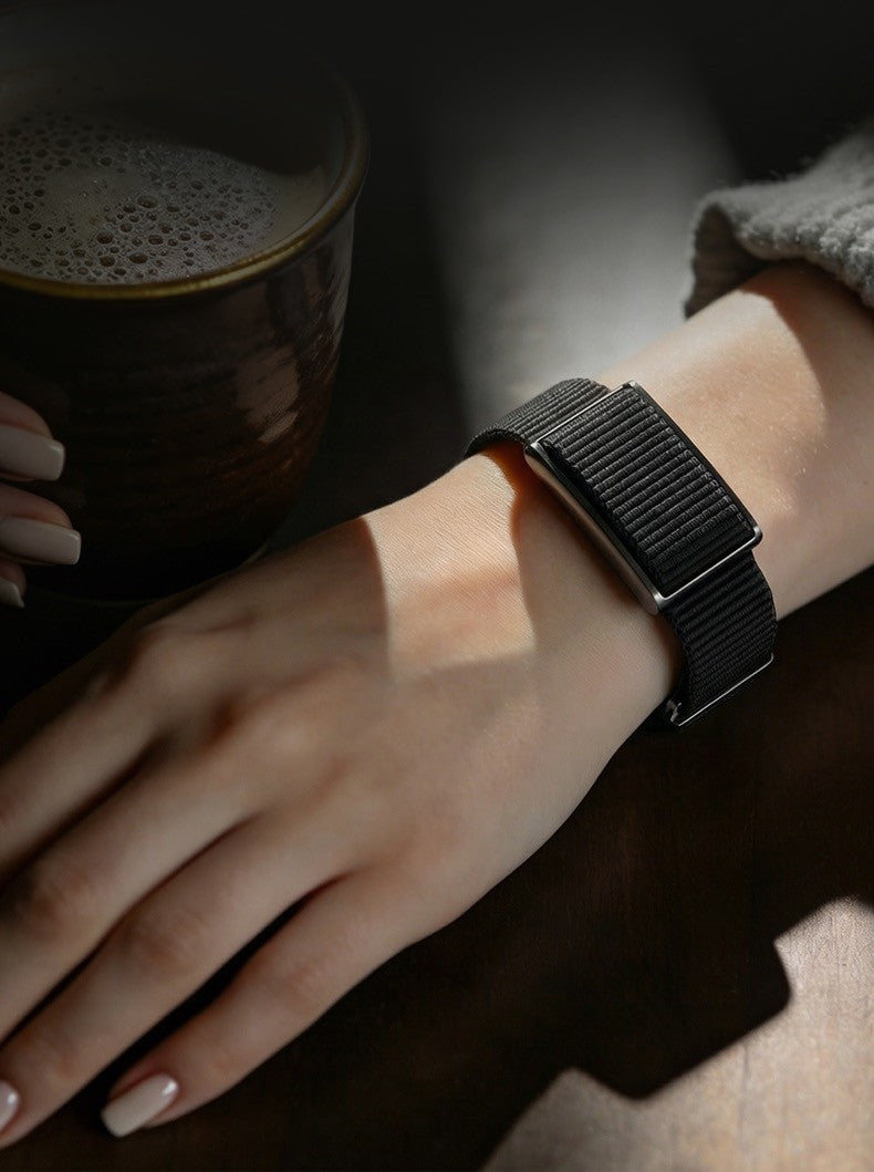 A close-up lifestyle shot of a person's hand and wrist wearing a black smartwatch with a textured black mesh band, resting near a dark ceramic mug on a dark wooden surface.